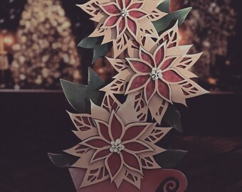 Może przedstawiać: Kompozycja papierowych kwiatów poinsecji w różowym wazonie z brązową podstawą. Kwiaty są warstwowe i mają efekt 3D. Kompozycja jest umieszczona na rozmytym tle choinki.