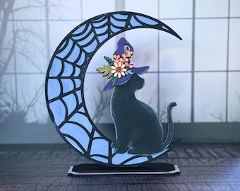 May include: A black cat wearing a purple witch hat with floral accents, perched on a blue crescent moon with a spiderweb design. The cat sits on a black base, perfect for Halloween decor.