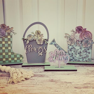 Puede incluir: Tres decoraciones de madera con temática primaveral. Una bota de lluvia a cuadros verde y blanco, un cubo gris con las palabras "flowers 5¢" y una regadera azul con las palabras "hello spring".