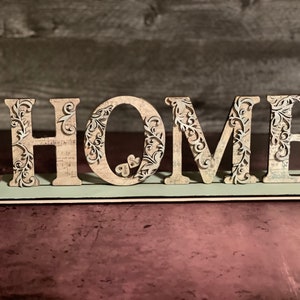 Może przedstawiać: Dekoracyjny drewniany znak z napisem "HOME" wielkimi literami. Litery są beżowe z misternymi kwiatowymi wycięciami i osadzone na jasnozielonej podstawie. Tło to rozmyta drewniana ściana.