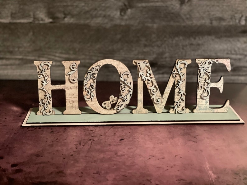 Może przedstawiać: Dekoracyjny drewniany znak z napisem "HOME" wielkimi literami. Litery są beżowe z misternymi kwiatowymi wycięciami i osadzone na jasnozielonej podstawie. Tło to rozmyta drewniana ściana.
