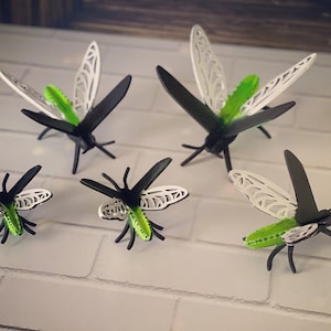 May include: Five black and white laser cut wood insects with green accents. The insects are in the shape of a dragonfly and are arranged on a white brick wall.