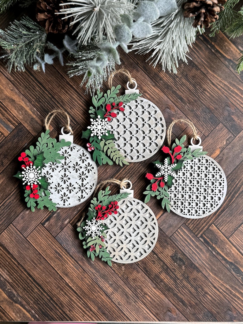 May include: Four white wooden Christmas ornaments with a geometric pattern and green and red paper foliage accents.