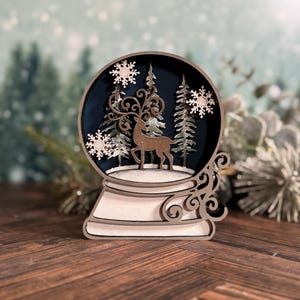 Puede incluir: Decoraci&oacute;n de globo de nieve de madera con un ciervo, &aacute;rboles y copos de nieve. El globo tiene un interior azul oscuro y se asienta sobre una base blanca con remolinos decorativos. La combinaci&oacute;n de colores es marr&oacute;n, blanco y azul.