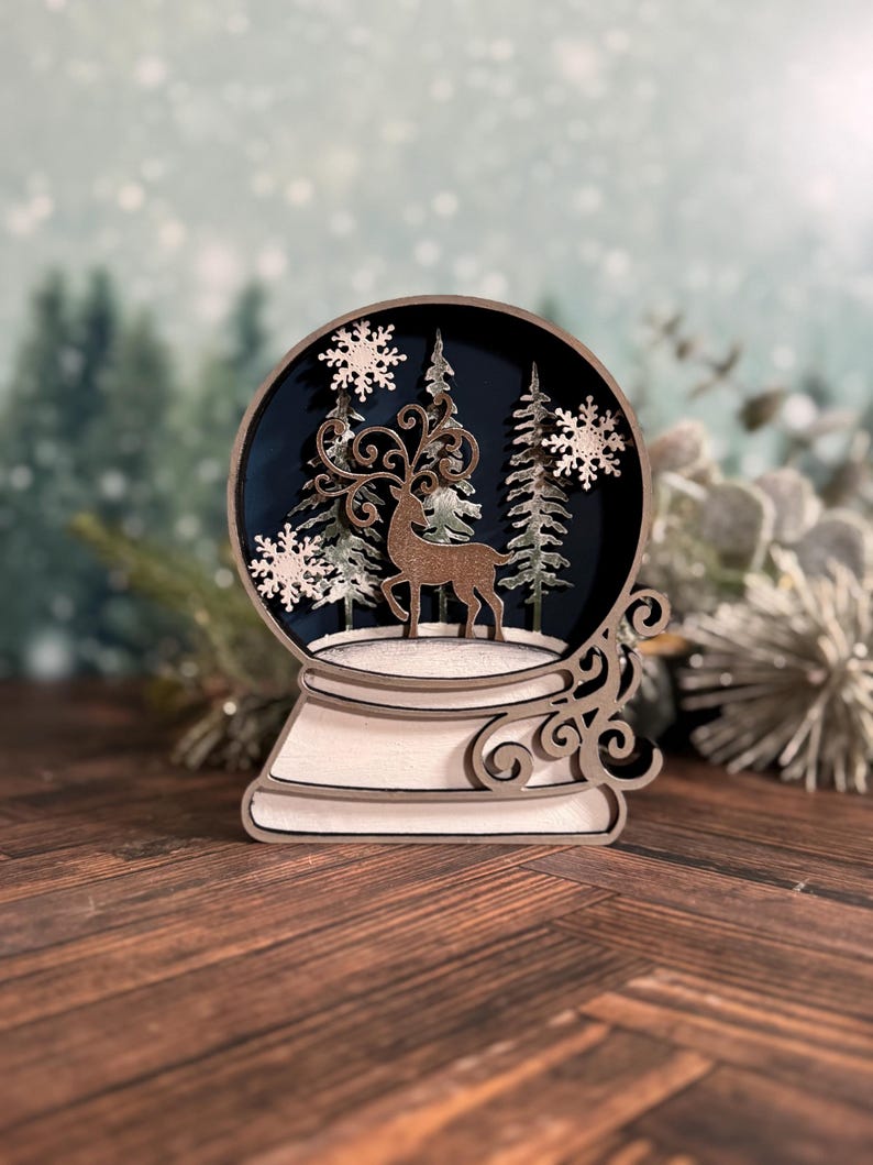 Puede incluir: Decoraci&oacute;n de globo de nieve de madera con un ciervo, &aacute;rboles y copos de nieve. El globo tiene un interior azul oscuro y se asienta sobre una base blanca con remolinos decorativos. La combinaci&oacute;n de colores es marr&oacute;n, blanco y azul.