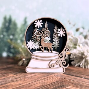 Puede incluir: Un globo de nieve decorativo con una escena invernal. El globo presenta un ciervo, &aacute;rboles y copos de nieve. La base y los detalles decorativos son de color claro, mientras que la escena interior es azul oscuro. Es una decoraci&oacute;n festiva y de temporada.