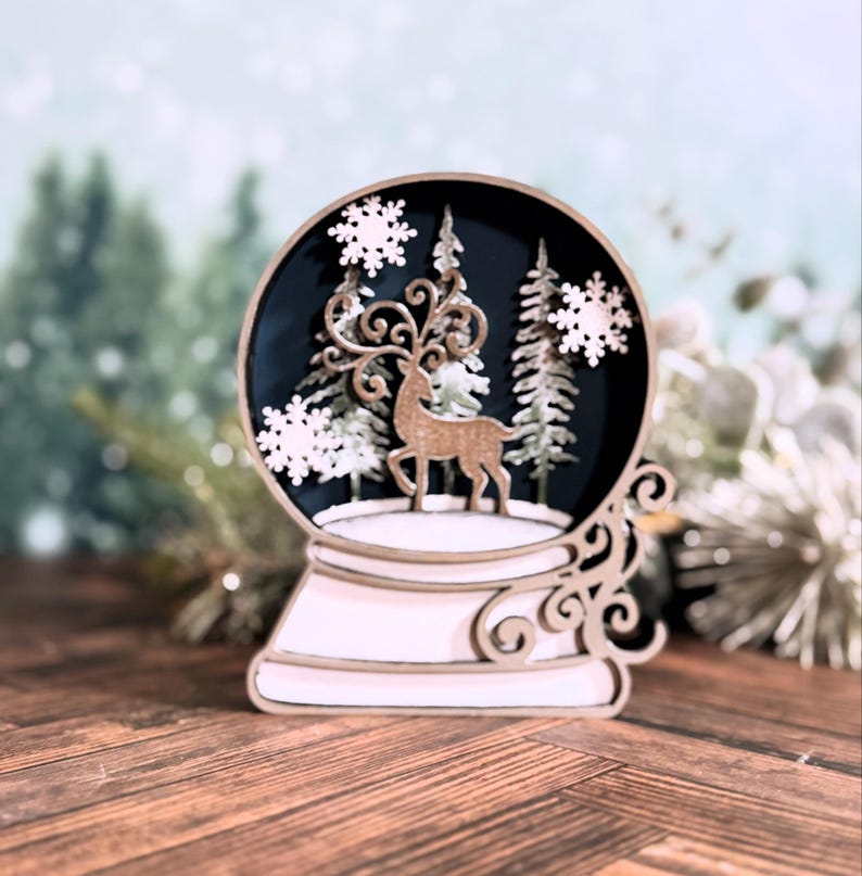 Puede incluir: Un globo de nieve decorativo con una escena invernal. El globo presenta un ciervo, &aacute;rboles y copos de nieve. La base y los detalles decorativos son de color claro, mientras que la escena interior es azul oscuro. Es una decoraci&oacute;n festiva y de temporada.