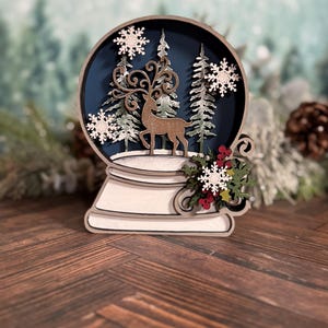 Puede incluir: Decoraci&oacute;n de globo de nieve de madera con un ciervo, &aacute;rboles y copos de nieve dentro de una esfera azul. La base es blanca y el dise&ntilde;o incluye bayas rojas y hojas verdes. El tema general es invierno y Navidad.