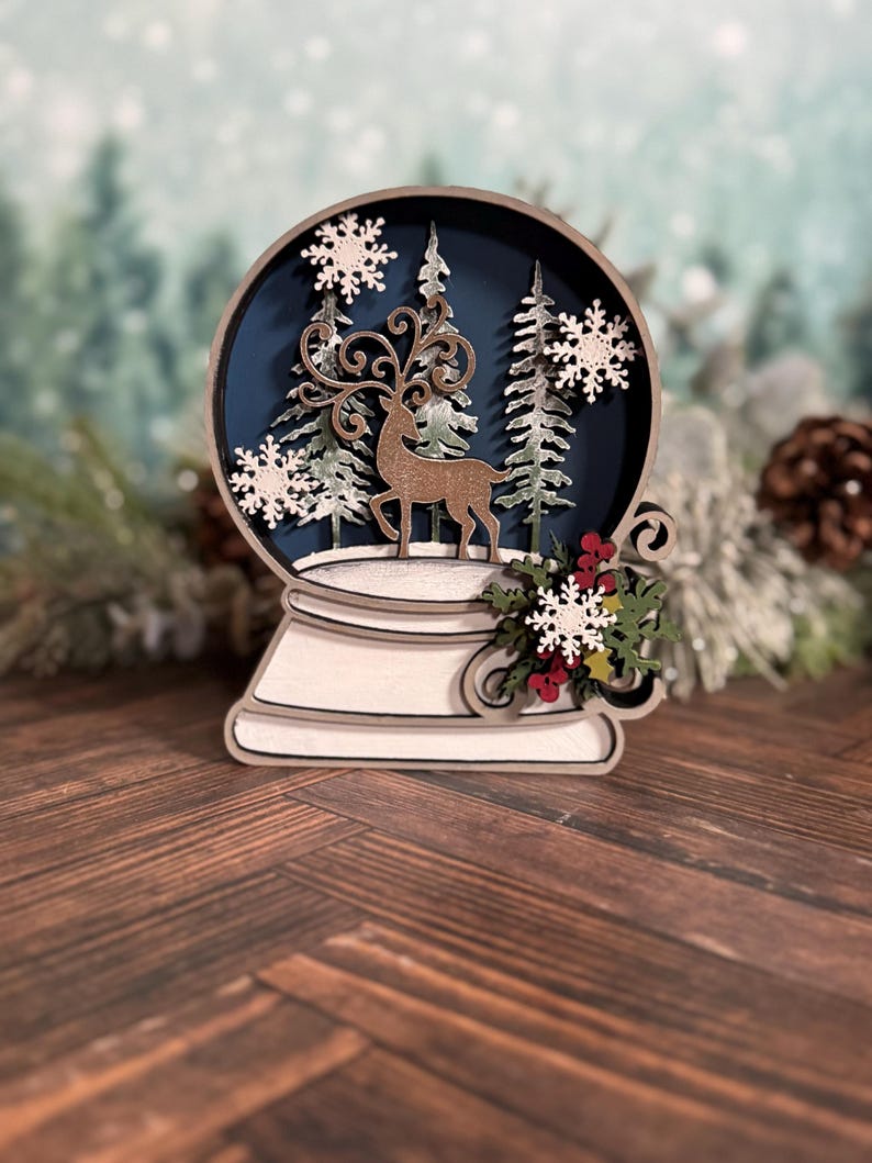 Puede incluir: Decoraci&oacute;n de globo de nieve de madera con un ciervo, &aacute;rboles y copos de nieve dentro de una esfera azul. La base es blanca y el dise&ntilde;o incluye bayas rojas y hojas verdes. El tema general es invierno y Navidad.