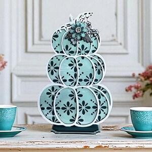 May include: A decorative, stacked pumpkin with a teal and white color scheme. The pumpkin has a floral design and is topped with a flower and leaf embellishment. The pumpkin is approximately 10 inches tall.