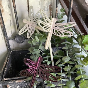 May include: Three decorative dragonfly garden stakes, two are white and one is a deep purple. The stakes are placed in a planter with green foliage.