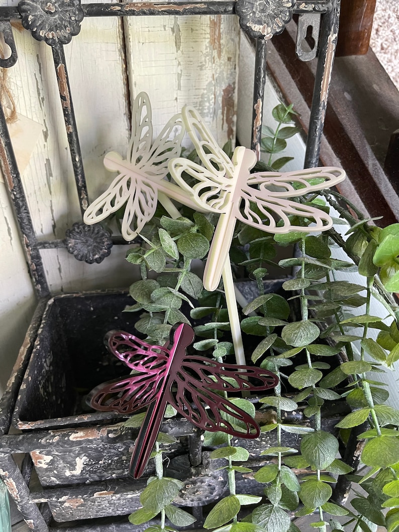 May include: Three decorative dragonfly garden stakes, two are white and one is a deep purple. The stakes are placed in a planter with green foliage.