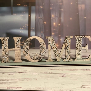 Może przedstawiać: Drewniany znak "HOME" z ozdobnymi, stylizowanymi literami. Jasne litery mają kwiatowe wzory i wycięcia. Prostokątna podstawa jest jasnozielona z czarno-białymi obramowaniami.