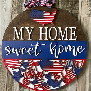 Può includere: Un cartello rotondo in legno con le parole "My Home Sweet Home" in lettere bianche. Il cartello è decorato con stelle e strisce rosse, bianche e blu, e un ritaglio degli Stati Uniti d'America.