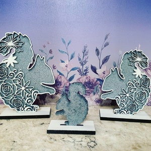May include: Three grey wooden squirrel silhouettes with floral designs. The squirrels are standing on separate white bases with black trim.