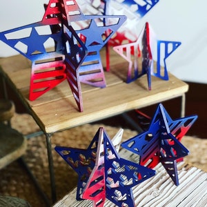 May include: Five wooden stars painted in red, white and blue with a layered, 3D design. The stars are arranged on two wooden surfaces.
