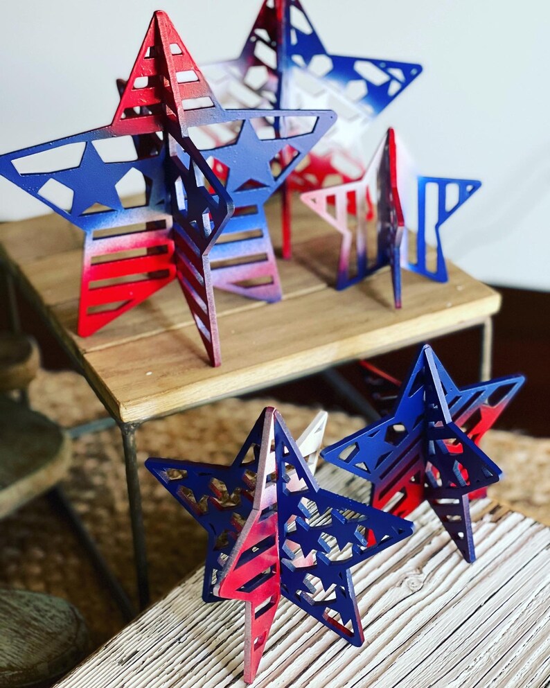 May include: Five wooden stars painted in red, white and blue with a layered, 3D design. The stars are arranged on two wooden surfaces.