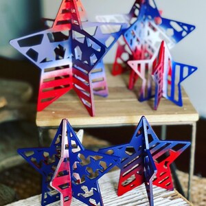 May include: Five red, white and blue wooden stars with a cutout design. The stars are painted with a gradient of red, white and blue.