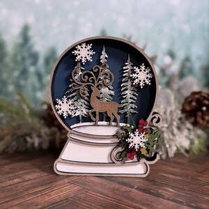 Puede incluir: Decoraci&oacute;n de globo de nieve de madera con un ciervo, &aacute;rboles y copos de nieve. El globo tiene un fondo azul oscuro y una base blanca. Incluye bayas rojas decorativas y hojas verdes. Adorno navide&ntilde;o.