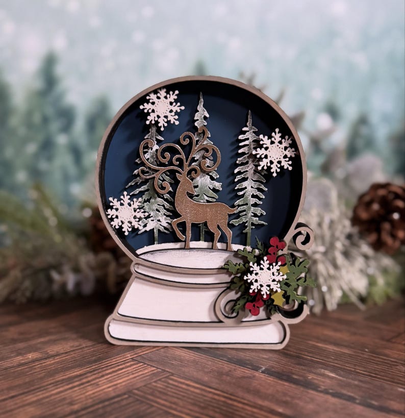 Puede incluir: Decoraci&oacute;n de globo de nieve de madera con un ciervo, &aacute;rboles y copos de nieve. El globo tiene un fondo azul oscuro y una base blanca. Incluye bayas rojas decorativas y hojas verdes. Adorno navide&ntilde;o.