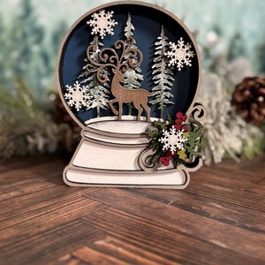 Puede incluir: Decoraci&oacute;n de globo de nieve de madera con un ciervo, copos de nieve y pinos dentro de una esfera azul. La base es blanca con remolinos decorativos y una ramita de bayas rojas y copos de nieve blancos. Tema invernal festivo.