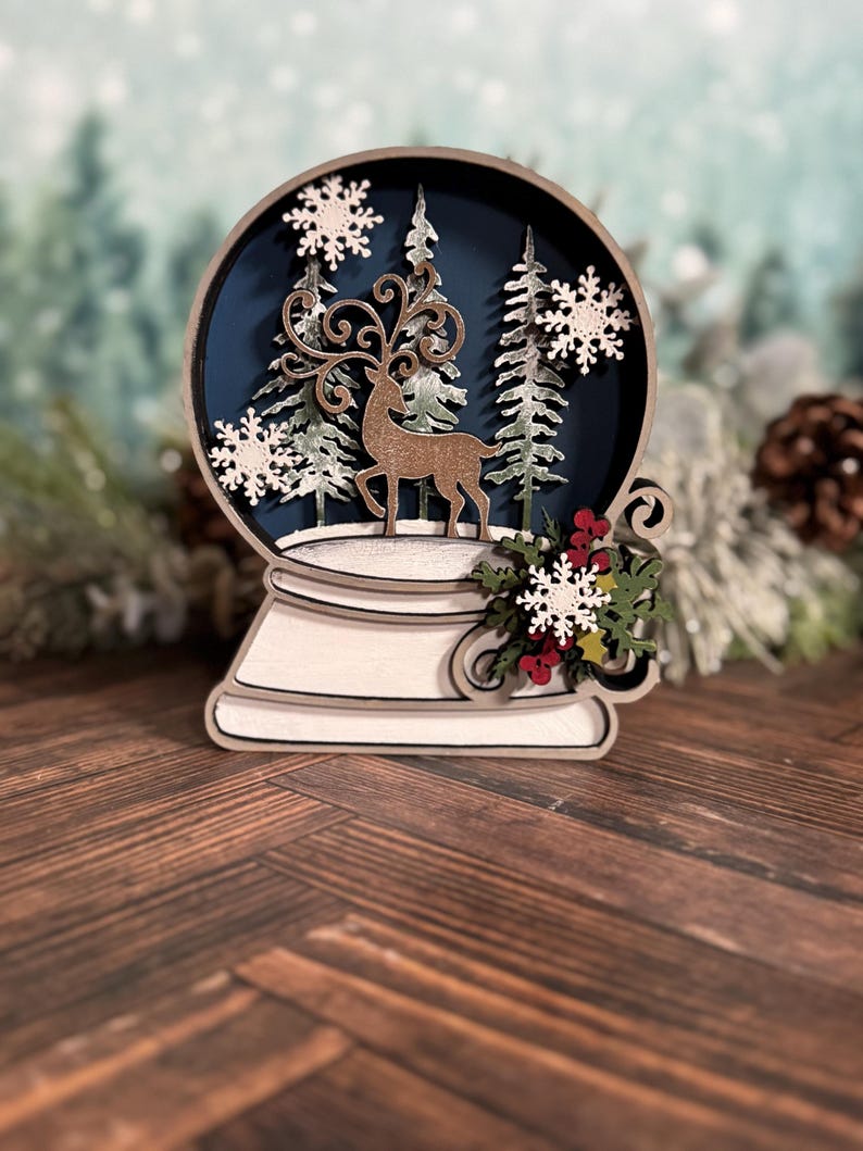 Puede incluir: Decoraci&oacute;n de globo de nieve de madera con un ciervo, copos de nieve y pinos dentro de una esfera azul. La base es blanca con remolinos decorativos y una ramita de bayas rojas y copos de nieve blancos. Tema invernal festivo.