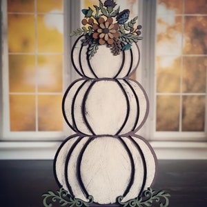 Puede incluir: Decoración de calabaza de tres niveles, blanca con contornos marrón oscuro y un arreglo floral en la parte superior. Las calabazas tienen un aspecto envejecido y rústico. El arreglo floral incluye acentos marrones, verdes y azules. Un adorno verde en la base.