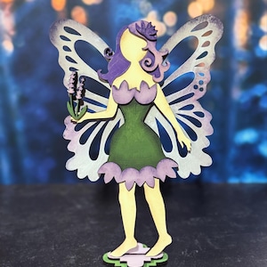 May include: A wooden fairy figurine with purple hair, green dress, and lavender flowers. The fairy has butterfly wings and stands on a pink and green base.