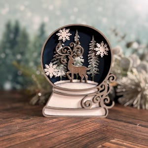 Puede incluir: Un globo de nieve de madera con una escena invernal. Dentro del globo, un reno se encuentra frente a &aacute;rboles y copos de nieve sobre un fondo azul oscuro. La base y los detalles decorativos son blancos y marrones.