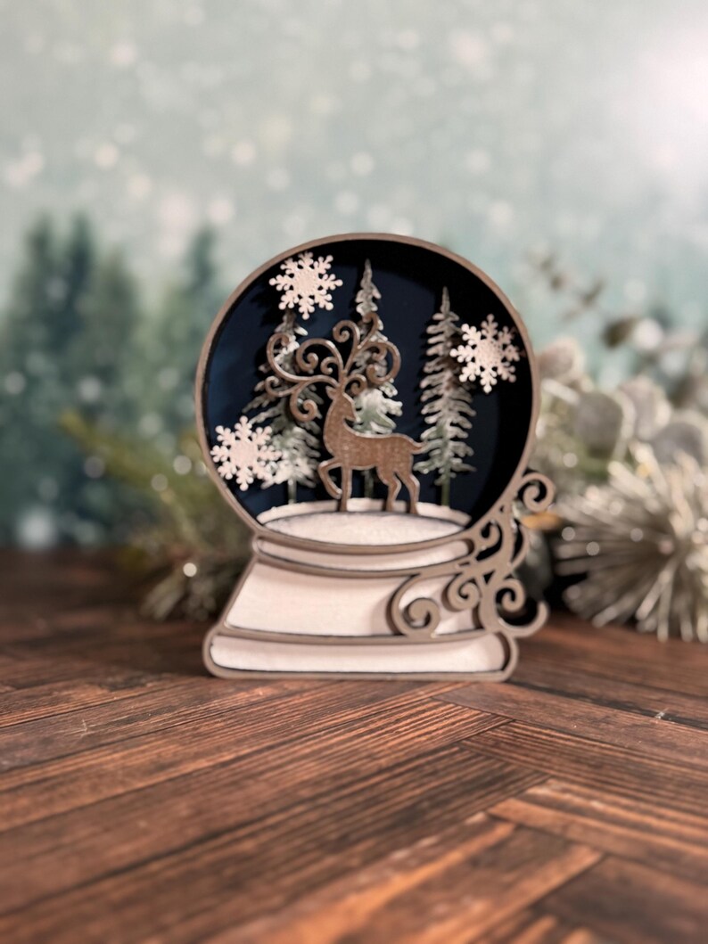 Puede incluir: Un globo de nieve de madera con una escena invernal. Dentro del globo, un reno se encuentra frente a &aacute;rboles y copos de nieve sobre un fondo azul oscuro. La base y los detalles decorativos son blancos y marrones.
