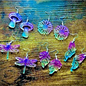 May include: A set of iridescent earrings featuring various nature-inspired designs, including mushrooms, sunflowers, dragonflies, butterflies, and monstera leaves.
