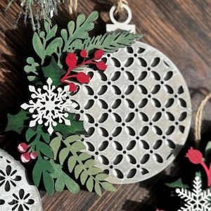May include: A set of three wooden Christmas ornaments with a white and gray geometric pattern. Each ornament is decorated with green leaves, red berries, and a white snowflake.