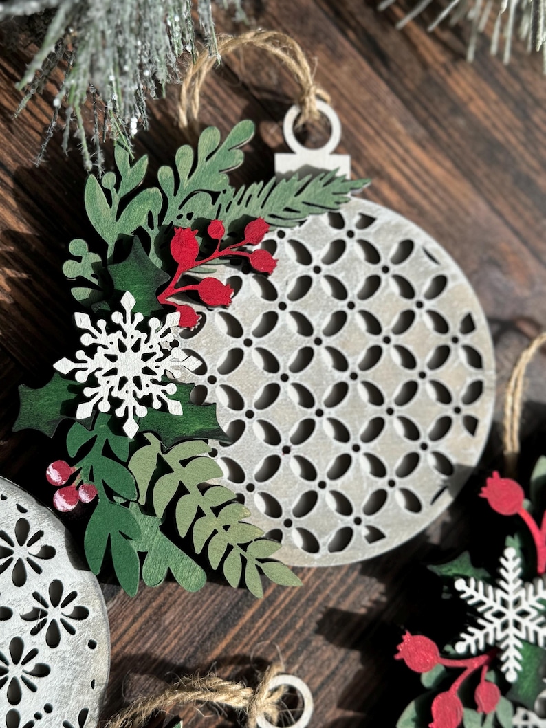 May include: A set of three wooden Christmas ornaments with a white and gray geometric pattern. Each ornament is decorated with green leaves, red berries, and a white snowflake.