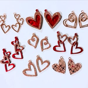 Puede incluir: Surtido de pendientes en forma de corazón en rojo y oro rosa. Algunos pendientes tienen brillo, mientras que otros tienen un diseño de espiral.