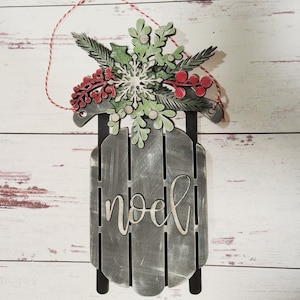 May include: A wooden Christmas sleigh ornament with a distressed gray finish. The sleigh is decorated with a green and white snowflake, red berries, and green leaves. The word "Noel" is written on the front of the sleigh.