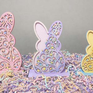 May include: Three decorative Easter bunny figurines in pastel colors. Each bunny has a layered design with intricate cut-out patterns. The bunnies are pink, purple, and yellow, and sit on a bed of colorful paper shreds.
