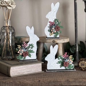 May include: Three white wooden bunny silhouettes with floral accents. Each bunny is decorated with a different arrangement of flowers in shades of pink, red, green, and yellow. The bunnies are displayed on a wooden surface.