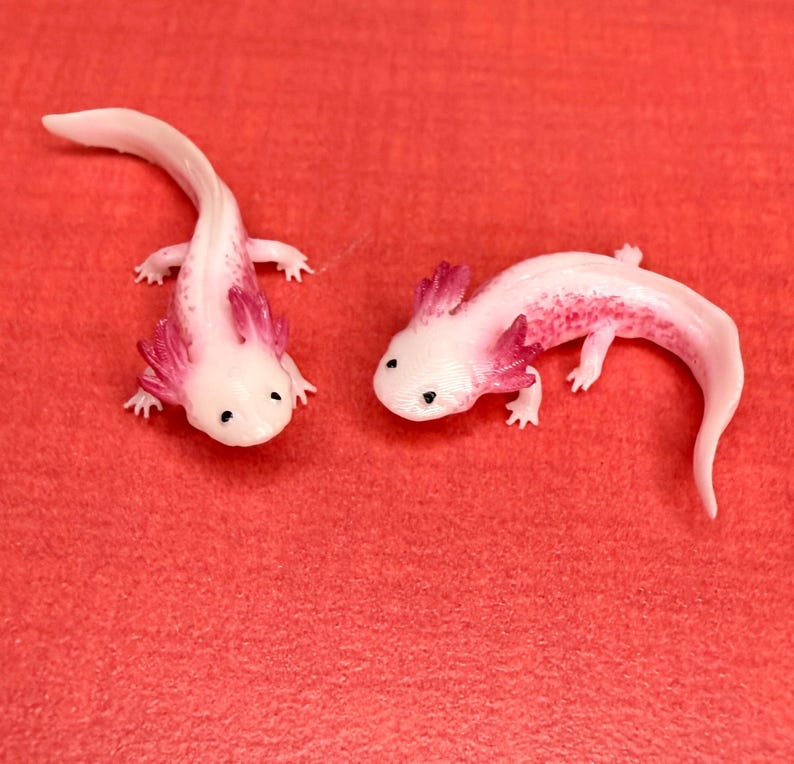 May include: Two pink and white axolotl figurines with red gills. The axolotls are posed on a red surface.