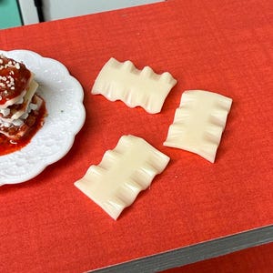 Puede incluir: Cuatro l&aacute;minas de lasa&ntilde;a de pl&aacute;stico blanco sobre una superficie roja. Las l&aacute;minas son ligeramente onduladas y tienen una superficie texturizada.