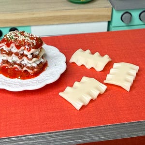 Puede incluir: Un plato blanco con un plato de lasa&ntilde;a en capas cubierto con salsa roja y hierbas verdes. Cuatro piezas de pasta de lasa&ntilde;a sin cocinar est&aacute;n sobre una superficie roja.