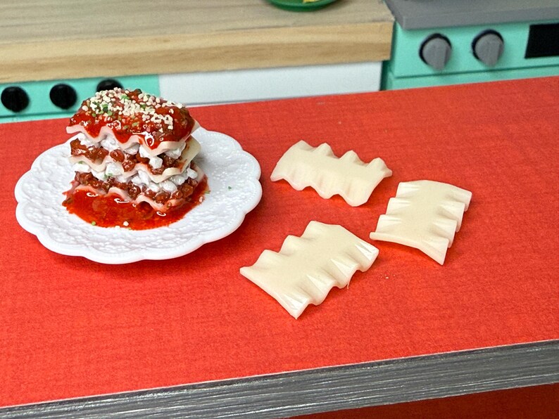Puede incluir: Un plato blanco con un plato de lasa&ntilde;a en capas cubierto con salsa roja y hierbas verdes. Cuatro piezas de pasta de lasa&ntilde;a sin cocinar est&aacute;n sobre una superficie roja.