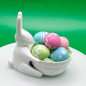 May include: A white ceramic bunny holding a basket filled with pastel colored Easter eggs. The eggs are decorated with stripes, dots, and swirls.