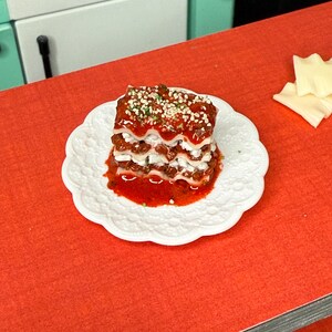 Puede incluir: Un peque&ntilde;o plato de lasa&ntilde;a cuadrado con tres capas de pasta, salsa de carne y queso. La lasa&ntilde;a est&aacute; cubierta con una pizca de hierbas verdes y una salsa roja.