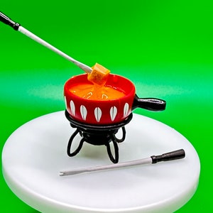 May include: A red and white ceramic fondue pot with a black metal stand and a long wooden handle dipping fork. The pot is filled with orange cheese fondue and a cube of bread is being dipped into the fondue.
