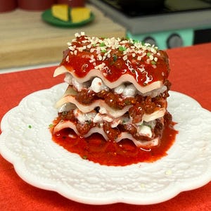 Puede incluir: Un plato blanco con una lasa&ntilde;a de tres capas cubierta con salsa roja y hierbas verdes. La lasa&ntilde;a est&aacute; sobre una superficie roja.