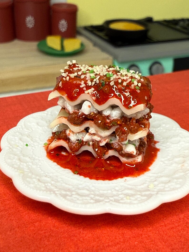 Puede incluir: Un plato blanco con una lasa&ntilde;a de tres capas cubierta con salsa roja y hierbas verdes. La lasa&ntilde;a est&aacute; sobre una superficie roja.