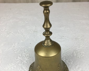 Pode incluir: Um sino de latão vintage com um padrão floral decorativo ao redor da base. O sino tem uma alça longa e um topo arredondado. O latão tem uma pátina quente e envelhecida. Este sino decorativo é adequado para decoração de casa.