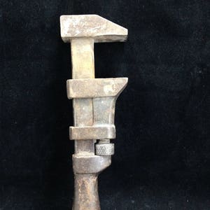 Puede incluir: Una llave inglesa ajustable vintage con cuerpo de metal y mango de madera. La llave tiene una cabeza en forma de T y un mecanismo de tornillo para ajustar el tamaño de la mordaza. La herramienta muestra signos de desgaste y edad, con una apariencia oxidada.