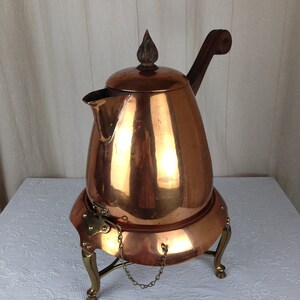 May include: A copper tea kettle with a wooden handle and lid finial. The kettle sits on a brass stand with curved legs and a small chain. The copper has a polished, reflective surface.
