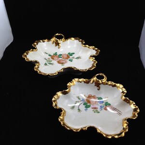 Könnte beinhalten: Zwei dekorative Schalen aus weißem Porzellan mit goldenen Verzierungen und Blumendesigns. Die Schalen haben einen gewellten Rand und einen Griff.